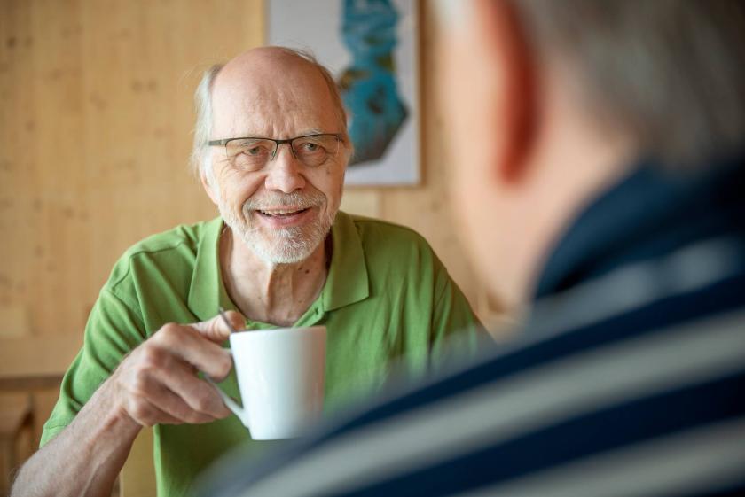 A man in a green shirt sitting and holding a coffee mug in one hand. I front of him there's another person who is only partly visible from behind.