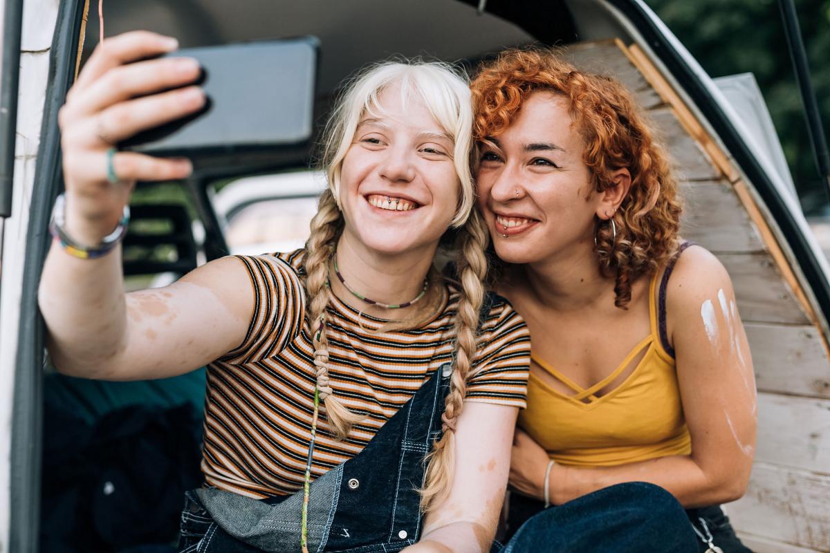 Kaksi naisoletettua istuvat ulkona ja ottavat itsestään selfietä kännykällä.